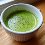 Close-up of vibrant green Cilantro Lime Crema with fine bubbles in a white bowl, sitting on a wooden surface.
