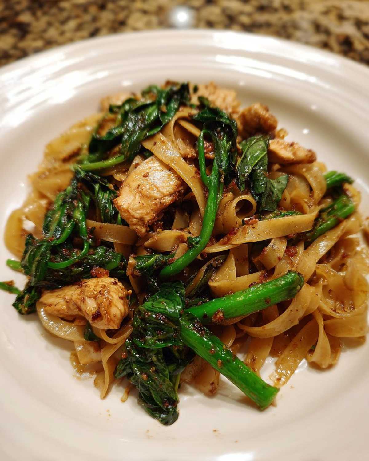 A close-up serving of Drunken Noodles Pad Kee Mao featuring wide rice noodles, pieces of chicken, and bright green Chinese broccoli.