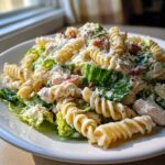 A close-up of Cold Chicken Caesar Pasta Salad featuring rotini pasta, romaine lettuce, chicken pieces, bacon bits, and grated Parmesan cheese.