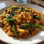 A close-up of a serving of Drunken Noodles Pad Kee Mao featuring wide rice noodles, green vegetables, and savory sauce on a white plate.