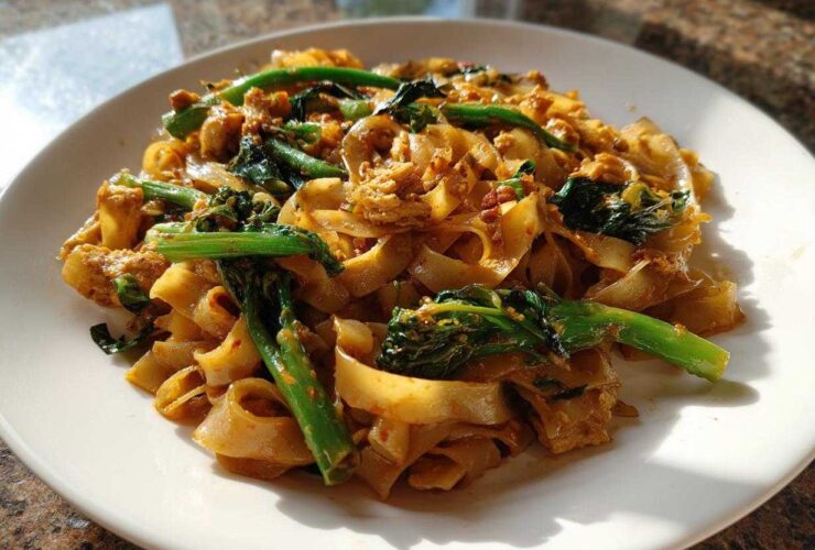 A close-up of a serving of Drunken Noodles Pad Kee Mao featuring wide rice noodles, green vegetables, and savory sauce on a white plate.
