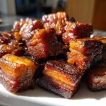 A close-up of glistening, caramelized Five-Spice Pork Belly Bites stacked on a white plate, catching the sunlight.