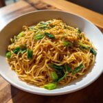 A close-up of a white bowl filled with glossy Ginger Scallion Noodles topped with bright green scallions.