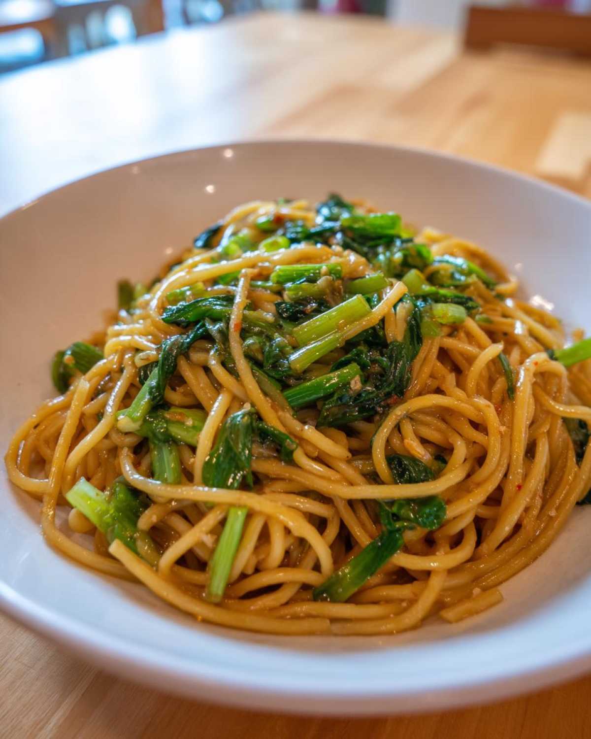 A close-up shot of a white bowl filled with glossy Ginger Scallion Noodles topped with vibrant green leafy vegetables.