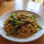 A white plate holding a generous serving of glossy Ginger Scallion Noodles mixed with bright green scallions.