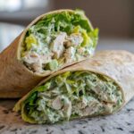 A close-up of Green Goddess Chicken Wraps cut in half, showing shredded chicken, lettuce, and creamy green dressing inside a whole wheat tortilla.