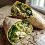 Two halves of a Green Goddess Chicken Wraps showing shredded chicken, lettuce, and creamy green dressing inside a whole wheat tortilla.