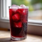 A tall, sweating glass filled with ice and deep red Agua Fresca de Jamaica Hibiscus, set near a bright window.