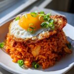 A mound of vibrant red Kimchi Fried Rice topped with a sunny-side-up crispy egg and green onions.