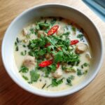 A close-up of a white bowl filled with creamy Lemongrass Coconut Chicken Soup, topped with fresh cilantro and sliced red chilies.