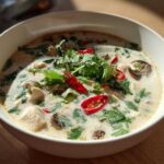 A close-up of a white bowl filled with creamy Lemongrass Coconut Chicken Soup, topped with cilantro and red chili slices.
