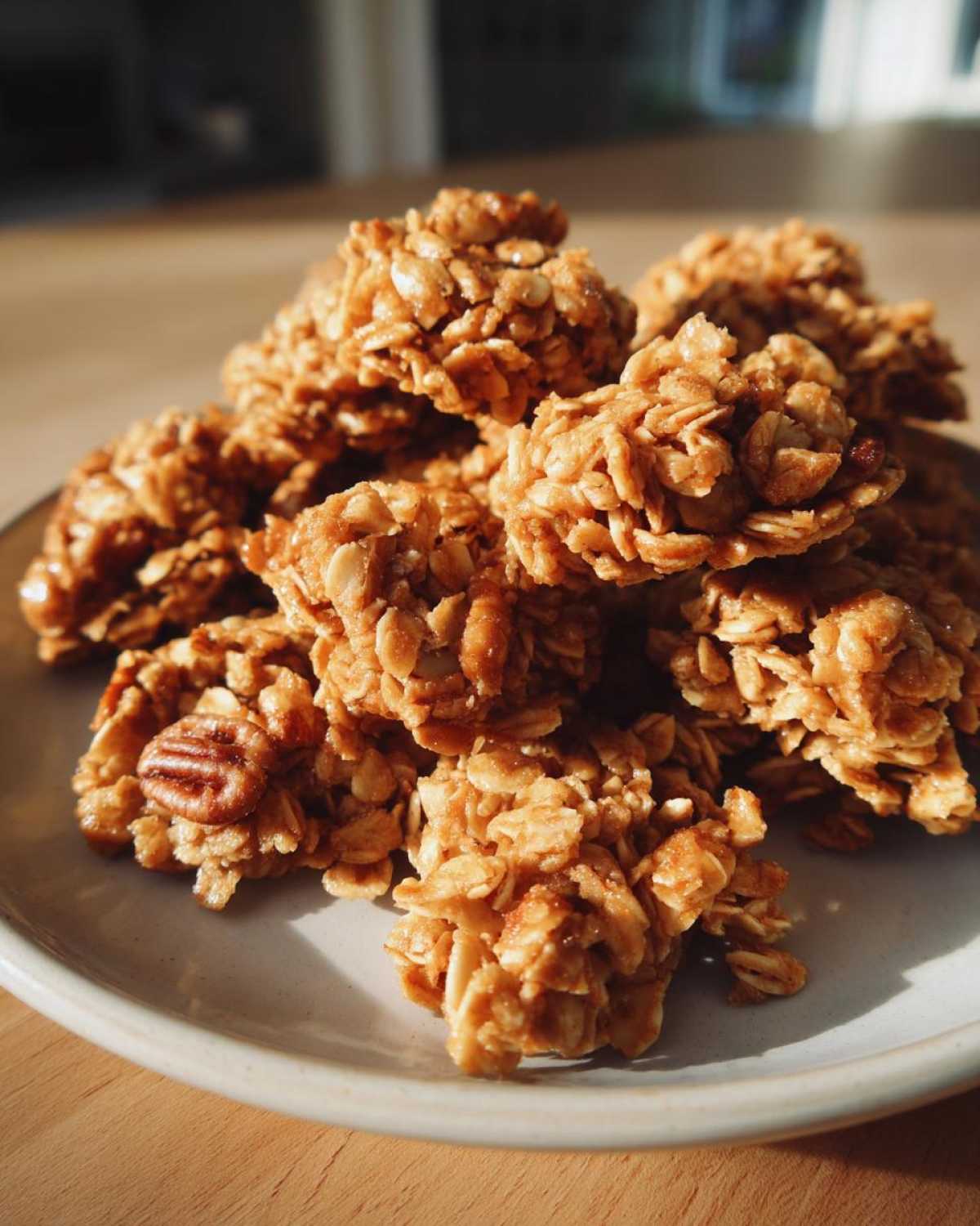 A pile of golden Maple Pecan Granola Clusters, featuring oats and visible pecans, resting on a light gray plate.