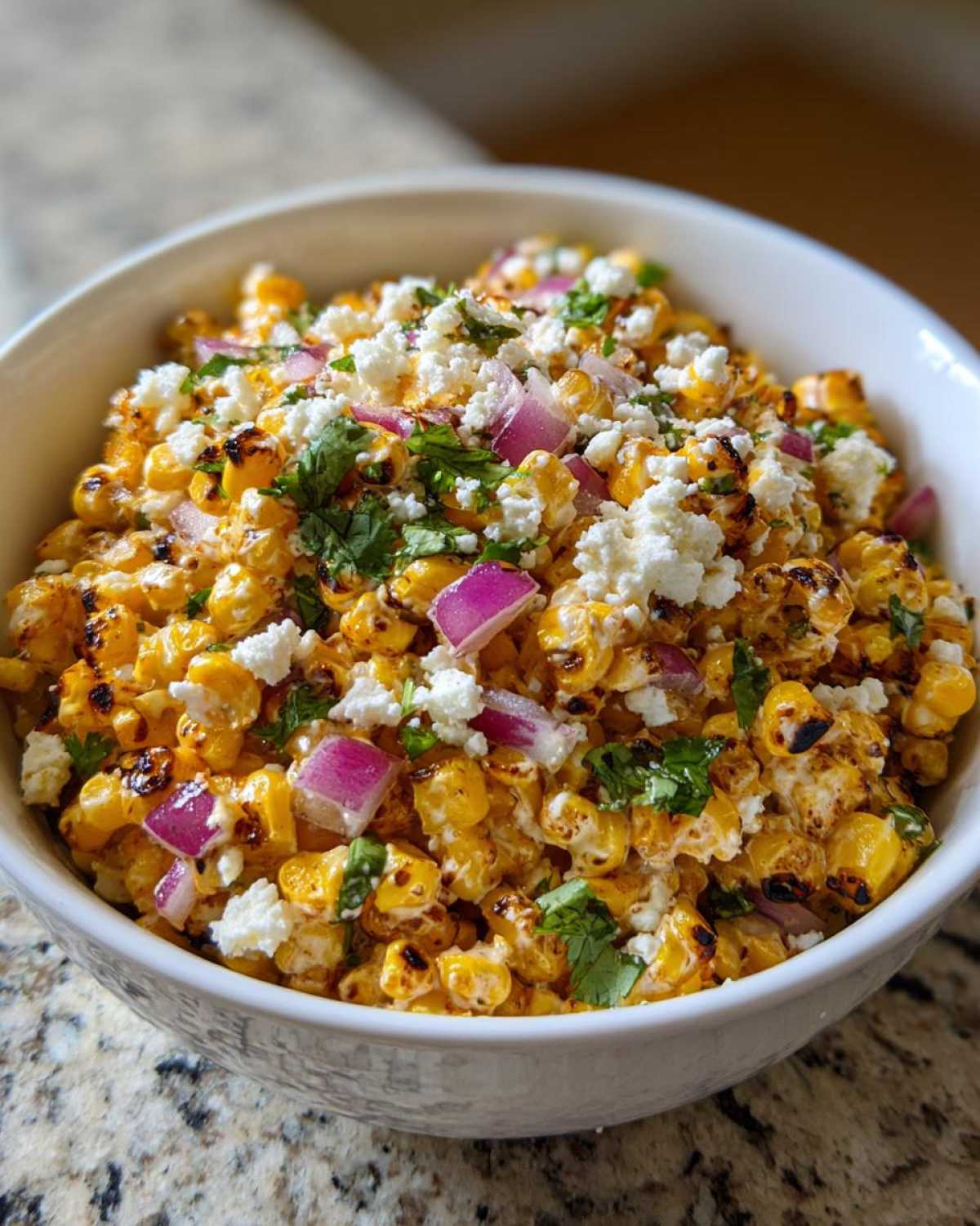 A close-up of a white bowl filled with Mexican Street Corn Salad Elote Style, topped with cotija cheese, red onion, and cilantro.