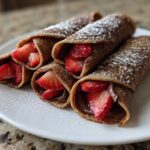 Four rolled Nutella Strawberry Crepe Rollups dusted with powdered sugar, served on a white plate.