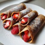 Six Nutella Strawberry Crepe Rollups dusted with powdered sugar, arranged on a white plate.