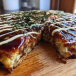 A thick, round Okonomiyaki Cabbage Pancake cut in half, drizzled with dark sauce and mayonnaise, topped with seaweed and bonito flakes.