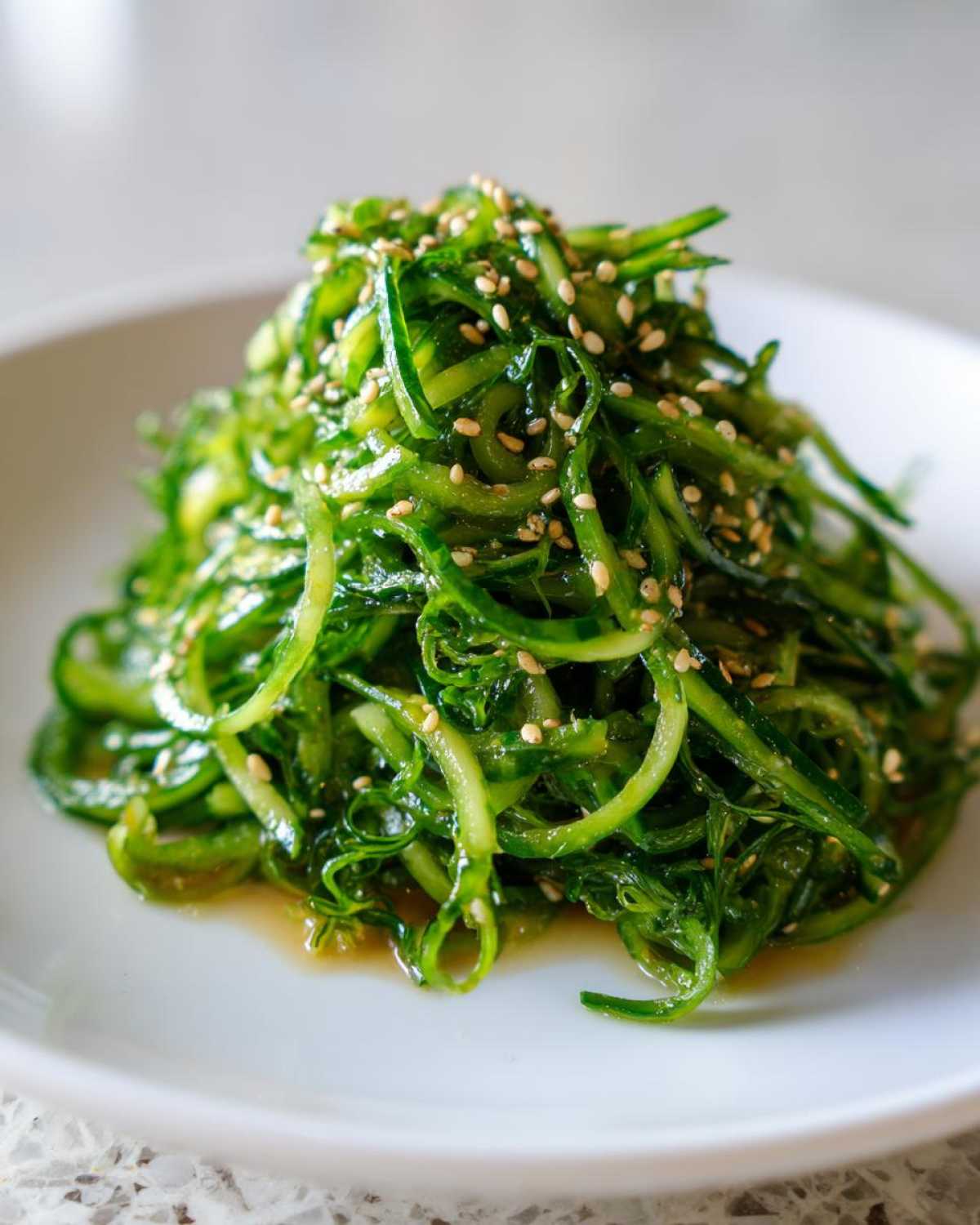 A mound of vibrant green, thinly sliced cucumber salad tossed in dressing and topped with toasted sesame seeds, resembling a Pea Shoot Cucumber Salad.