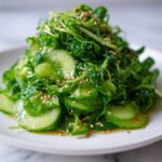 A vibrant mound of fresh Pea Shoot Cucumber Salad tossed in dressing and sprinkled with toasted sesame seeds, served on a white plate.