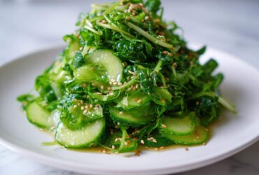 A vibrant mound of fresh Pea Shoot Cucumber Salad tossed in dressing and sprinkled with toasted sesame seeds, served on a white plate.