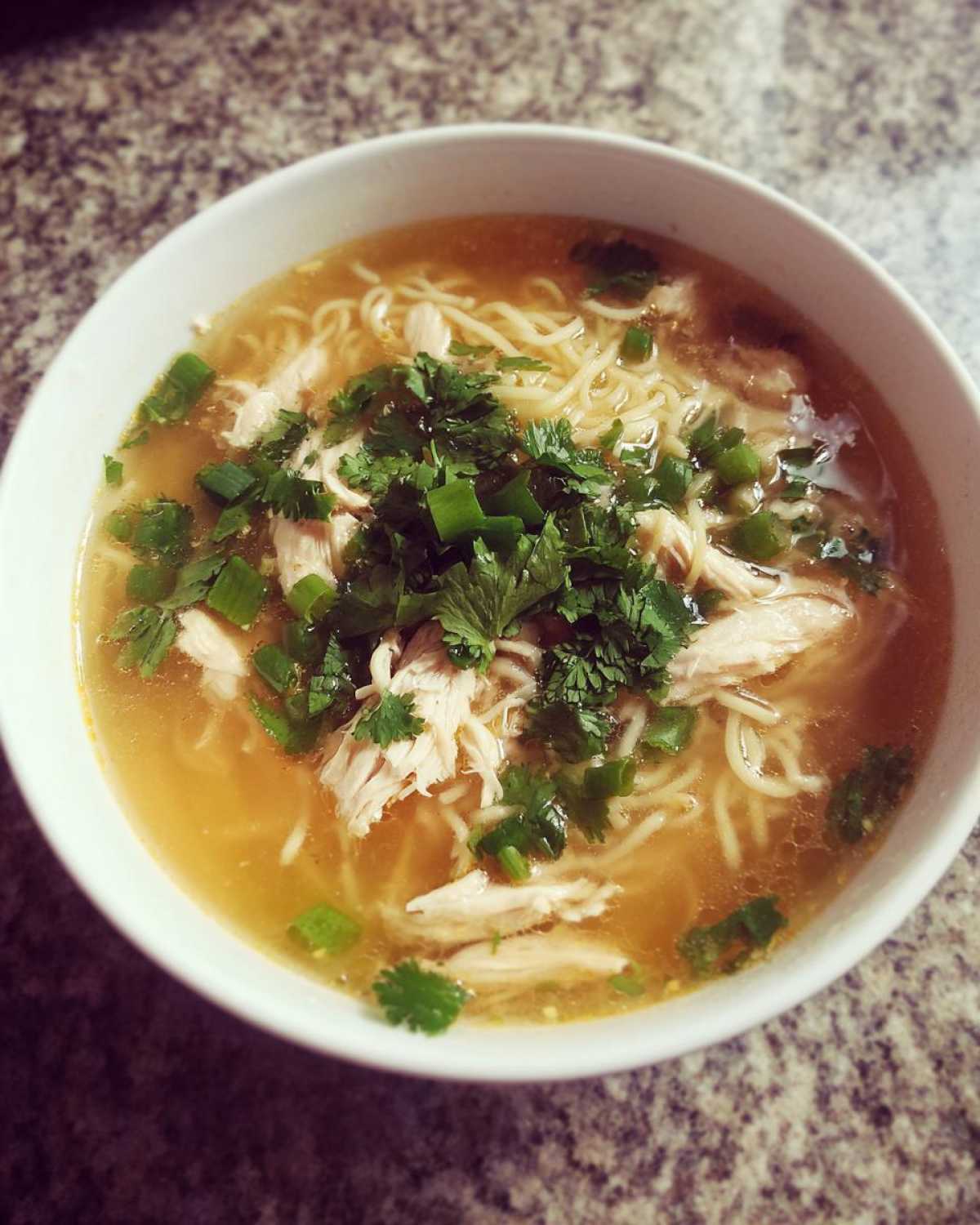 A close-up overhead view of a white bowl filled with Pho Ga Chicken Noodle Soup, topped with shredded chicken and fresh herbs.