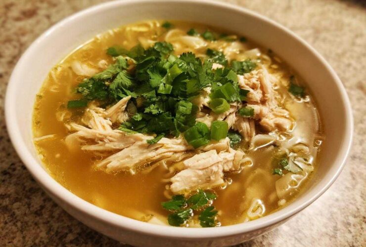 A close-up of a white bowl filled with rich broth, shredded chicken, noodles, and topped with fresh cilantro and scallions for Pho Ga Chicken Noodle Soup.