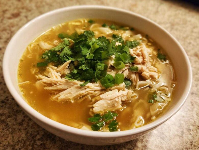 A close-up of a white bowl filled with rich broth, shredded chicken, noodles, and topped with fresh cilantro and scallions for Pho Ga Chicken Noodle Soup.