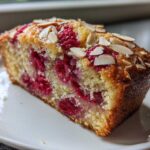 A close-up of a moist slice of Raspberry Almond Olive Oil Cake studded with bright red raspberries and topped with sliced almonds.