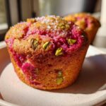 A close-up of a freshly baked Raspberry Pistachio Muffin topped with coarse sugar and green pistachio pieces.