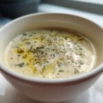 Close-up of creamy Roasted Garlic White Bean Soup topped with olive oil and herbs in a white bowl.