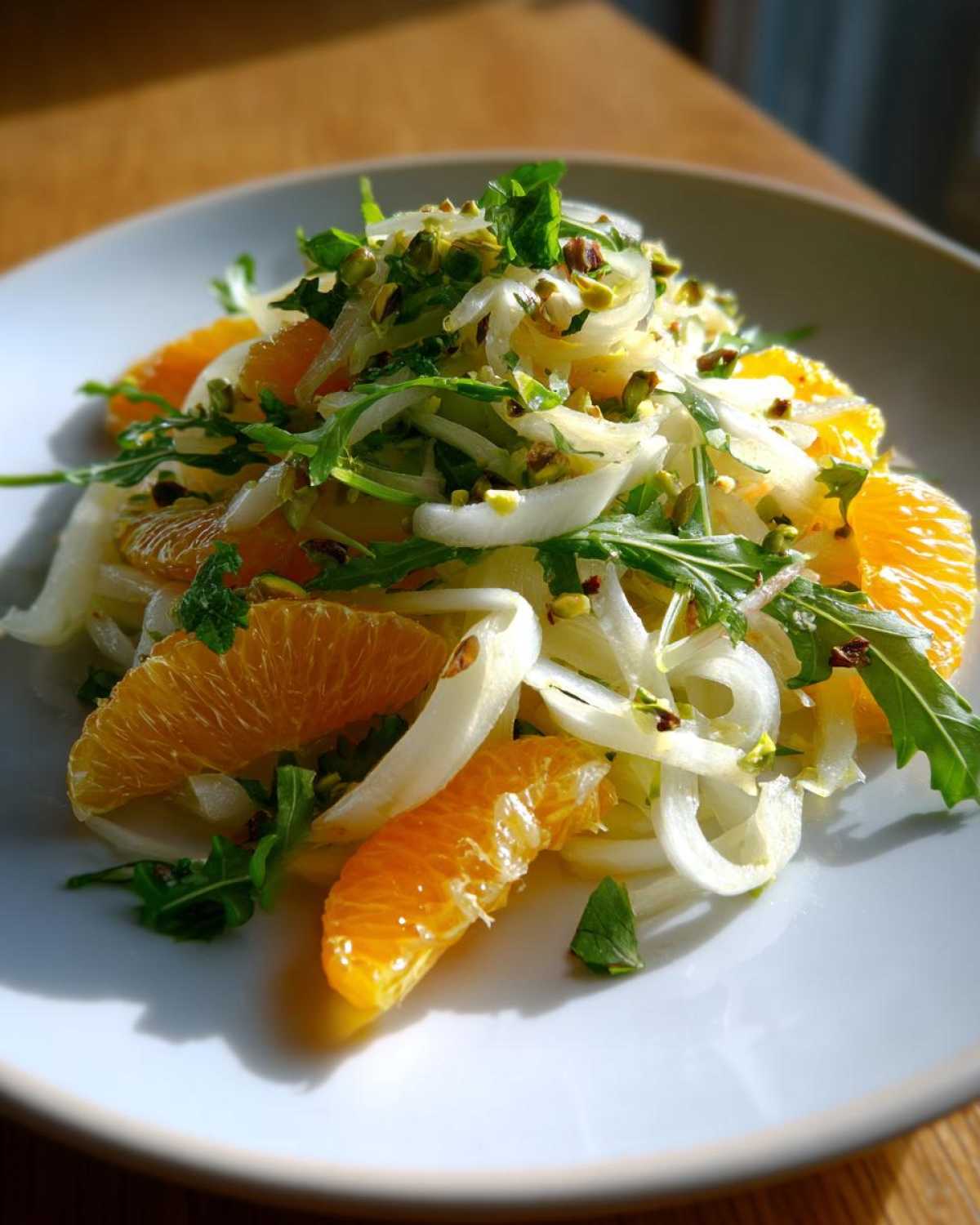 Close-up of a vibrant Shaved Fennel Citrus Salad featuring orange segments, arugula, and chopped pistachios on a white plate.
