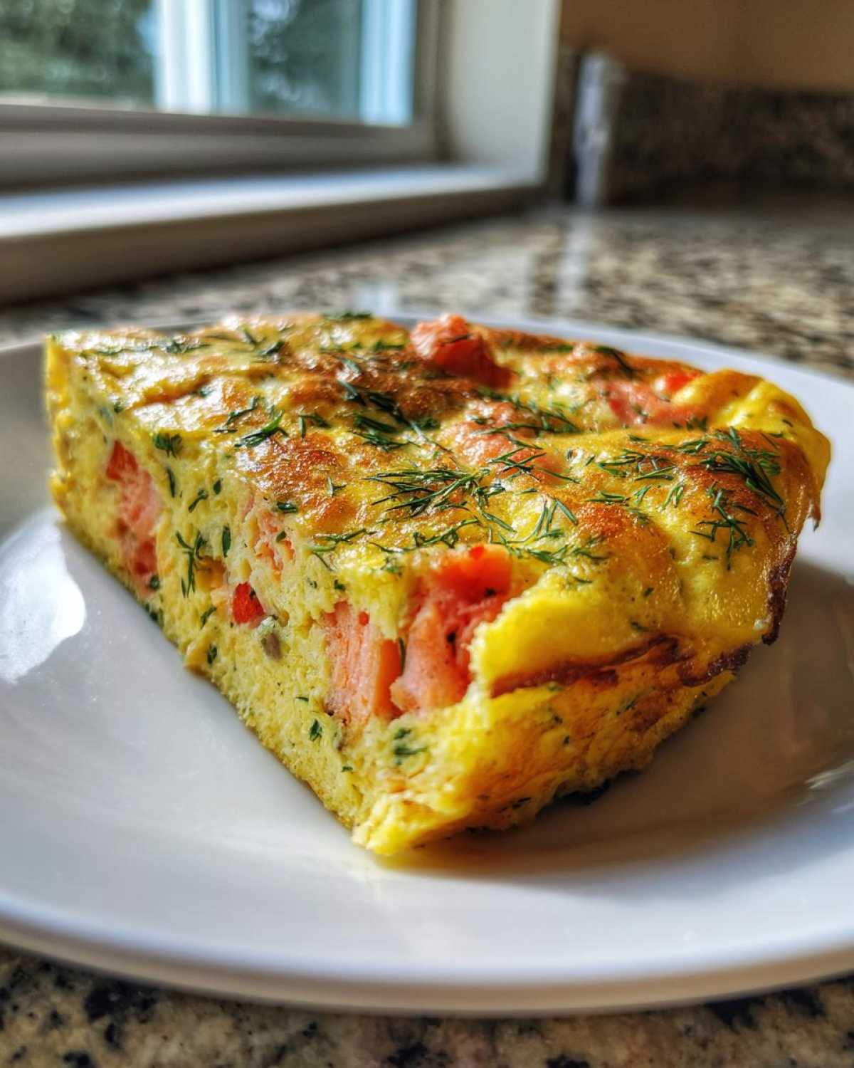 A thick, golden slice of Smoked Salmon Dill Frittata topped with fresh dill, served on a white plate.