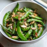 Close-up of a bright green Snap Pea Sesame Salad topped with toasted sliced almonds and sesame seeds.