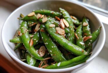 Close-up of a bright green Snap Pea Sesame Salad tossed with sesame seeds and sliced almonds in a white bowl.