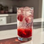 Close-up of a tall glass filled with Sparkling Strawberry Shrub Soda, ice, and whole berries, bubbling vigorously.