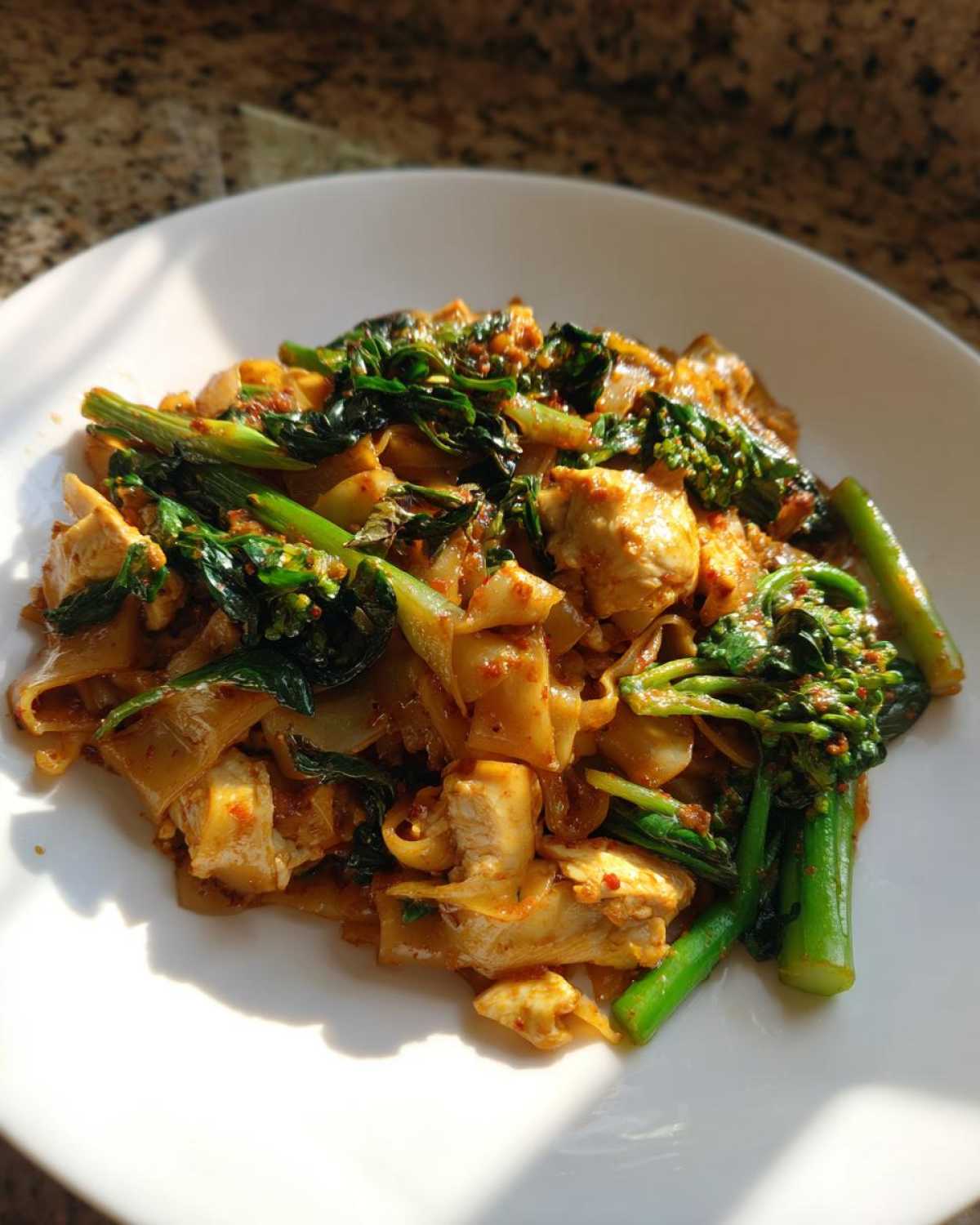 A plate of spicy Drunken Noodles Pad Kee Mao featuring wide rice noodles, chicken pieces, and bright green Chinese broccoli.