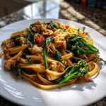 A close-up of a serving of Drunken Noodles Pad Kee Mao featuring wide noodles, chicken pieces, basil, and broccolini on a white plate.