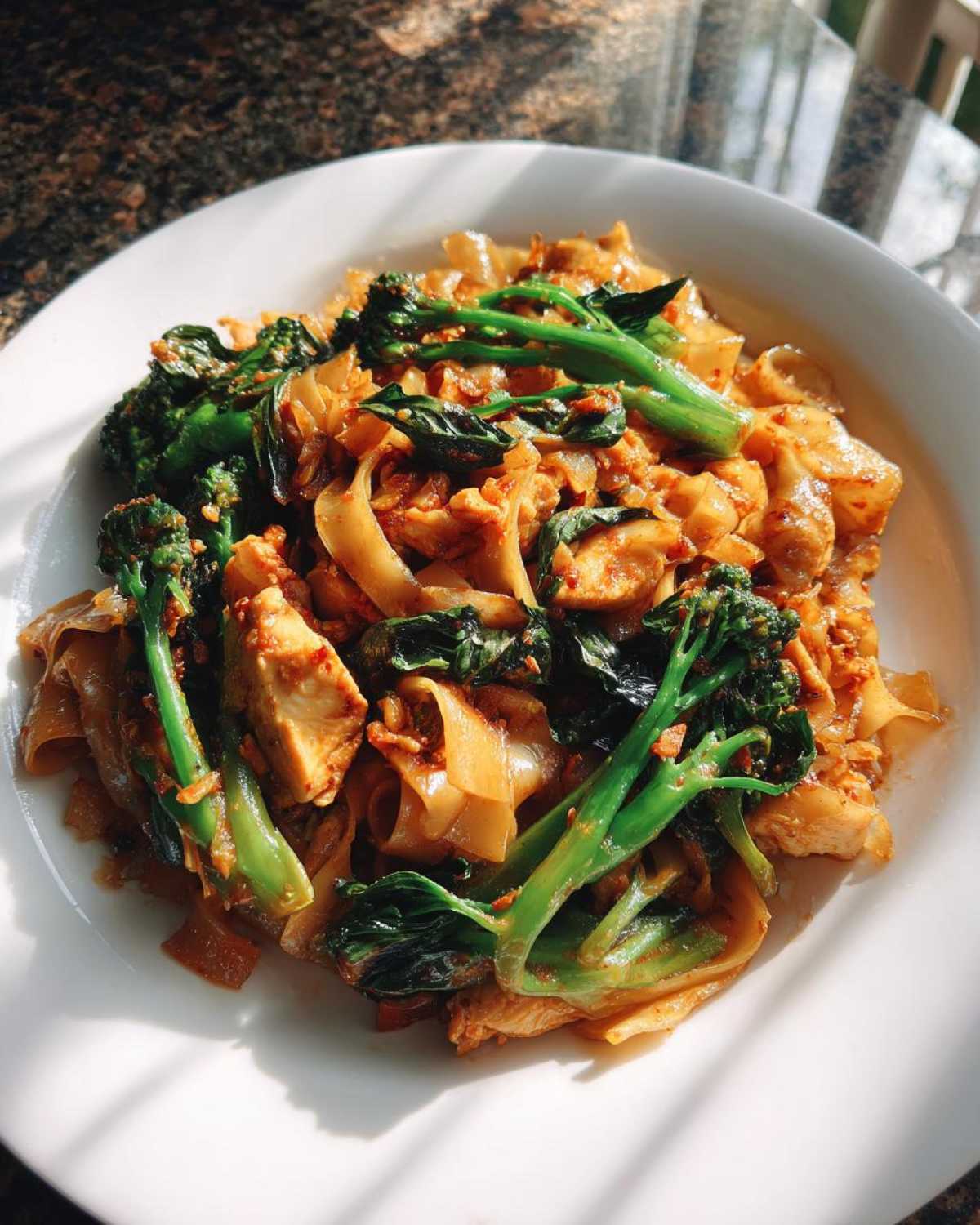 A close-up of a plate of spicy Drunken Noodles Pad Kee Mao featuring wide rice noodles, chicken pieces, and vibrant green Chinese broccoli.