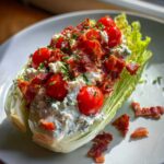 A close-up of a Steakhouse Wedge Salad with Blue Cheese dressing, topped with bacon bits, cherry tomatoes, and chives.