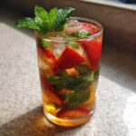 A tall, sweating glass filled with Strawberry Iced Green Tea with Mint, ice, sliced strawberries, and fresh mint leaves.
