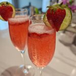 Two flutes filled with a fizzy, pink Strawberry Mimosa Mocktail, garnished with a fresh strawberry and lime slice.