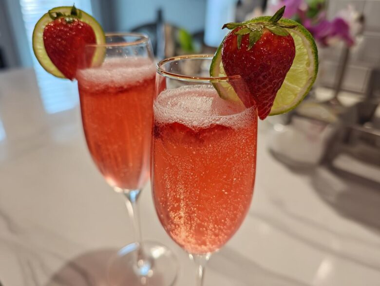 Two flutes filled with a fizzy, pink Strawberry Mimosa Mocktail, garnished with a fresh strawberry and lime slice.