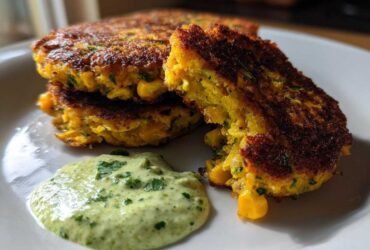 A stack of golden brown Sweet Corn Zucchini Fritters served with a dollop of creamy green herb sauce on a white plate.