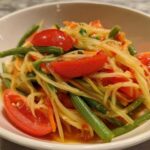 Close-up of freshly prepared Thai Papaya Salad Som Tam featuring shredded green papaya, bright red tomato wedges, and green beans.