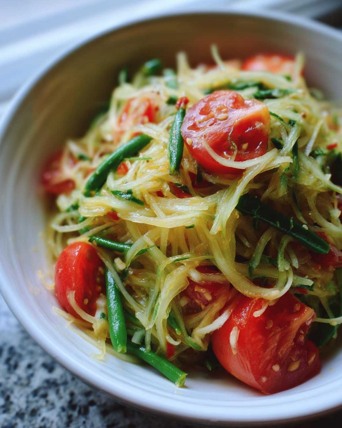 A close-up of vibrant Thai Papaya Salad Som Tam featuring shredded green papaya, cherry tomatoes, and green beans in a white bowl.