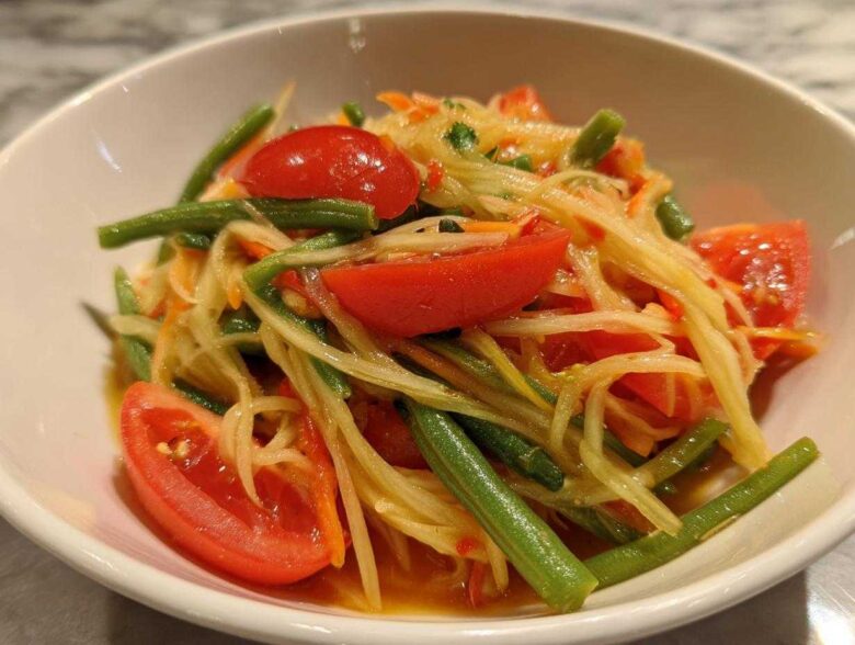 Close-up of freshly prepared Thai Papaya Salad Som Tam featuring shredded green papaya, bright red tomato wedges, and green beans.