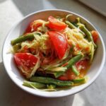 Close-up of vibrant Thai Papaya Salad Som Tam featuring shredded green papaya, bright red tomatoes, and green beans in a white bowl.