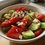 A close-up of a vibrant Tomato Cucumber Feta Salad with chunks of red tomato, green cucumber, and crumbled white feta, seasoned with herbs.