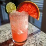 A tall glass filled with a fizzy, pink Virgin Paloma Grapefruit Spritz, garnished with a lime wheel and a slice of ruby red grapefruit.