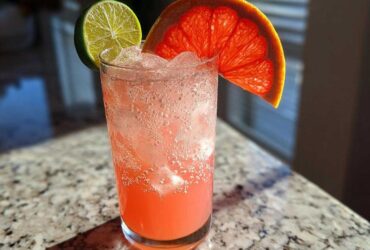 A tall glass filled with a fizzy, pink Virgin Paloma Grapefruit Spritz, garnished with a lime wheel and a slice of ruby red grapefruit.